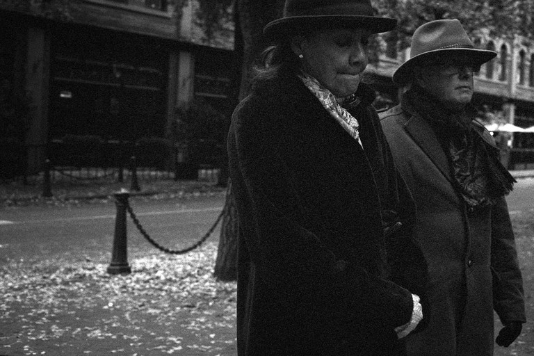 A couple in hats and overcoats pensively walk through a dimly lit street.