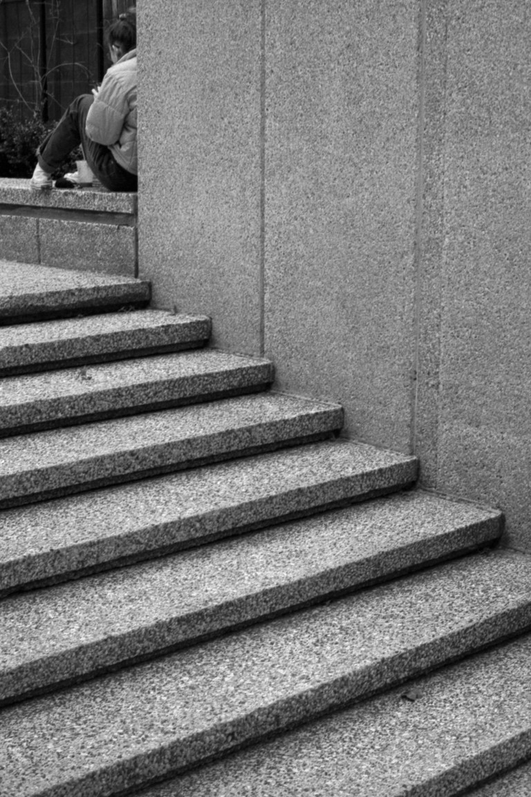 A girl sits nestled into a geometric corner of a brutalist concrete scene. 