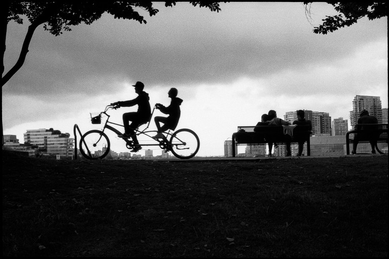 People in silhouette sit on benches in the park as two people on a bicycle-built-for-two roll through.