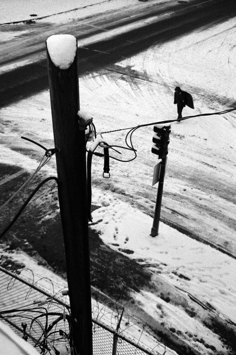 A man crosses an icy street. In the foreground, a belt hangs from an electrical pole.