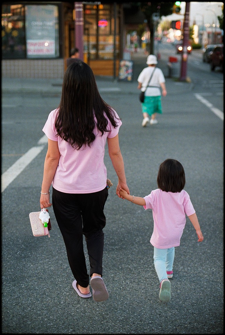 A mother and daughter in matching pink shirts cross the street on a summer evening.