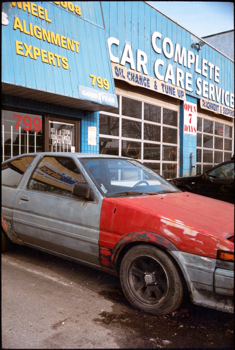 A weathered and garishly painted car sits in front of a similarly weathered and garish auto repair store front.