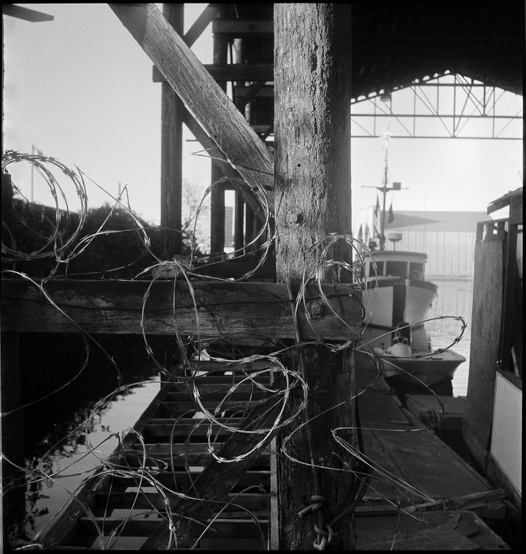An old boat is moored in a covered boathouse surrounded by barbed wire.