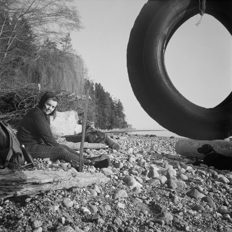 A woman and dog sit on a beach beneath a tire swing. The scale of the items in view is slightly compressed and hard to discern.