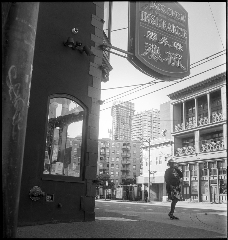 A woman enters an intersection surrounded by older buildings. The Vancouver skyline is in the distance.