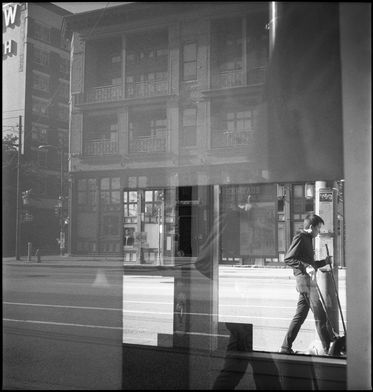 A man with broom and dustbin is reflected in a mirror on Pender Street in Chinatown.