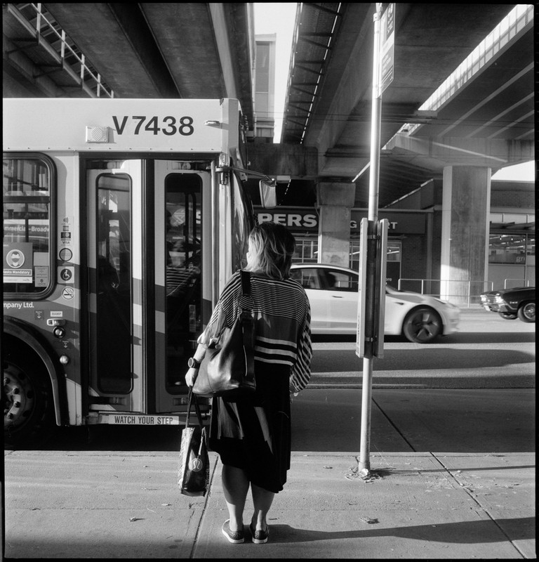 A woman waits for the bus, her striped top echoing the shadows and geometry around her.